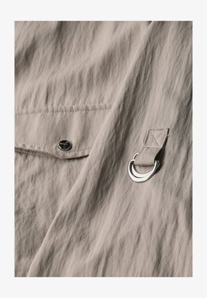 Nära bild av beige tyg med en texturerad yta, som har en svart tryckknapp och en silverfärgad metallring fäst med ett tygband.