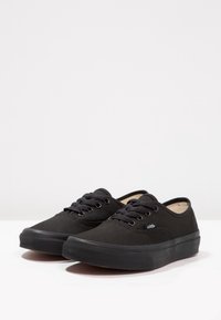 Un par de zapatillas Vans negras de corte bajo con cordones negros y suelas de goma sobre una superficie blanca.