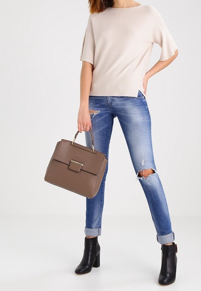 Top beige a coste; jeans skinny blu strappati; borsa in pelle marrone con chiusura; stivaletti neri con tacco; sfondo neutro.