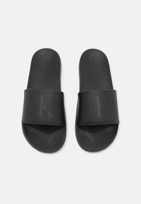 Sandales noires à semelles plates avec une large bride présentant un logo 'CK' en relief. Fabriquées en matériau synthétique lisse. Semelle intérieure plate et texturée.