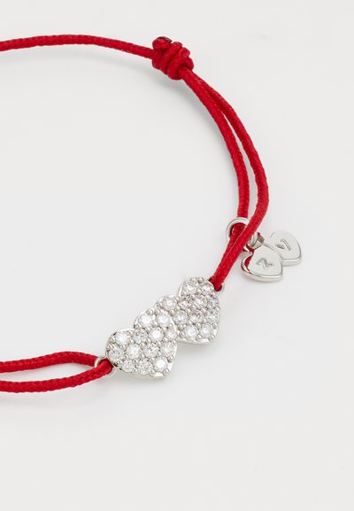 Red cord bracelet featuring two interconnected hearts, adorned with clear sparkling crystals. Silver accent with small heart tag attached.