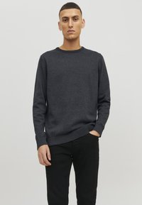 Suéter de punto gris oscuro con cuello redondo, mangas largas y puños y dobladillo acanalados. Llevado con pantalones negros, sobre un fondo claro.
