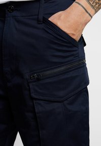 Navy cargo pants featuring a zippered pocket, button closure, and textured fabric. Includes side pockets with pronounced stitching details.