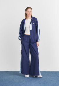 Merinavi sininen tuulihaalari, jossa on vaaleansinisiä yksityiskohtia, joustavat hihansuut ja vyötärö. Hölkköhousut merinavi sinisestä, joissa on sivutaskut ja Nike-logo.