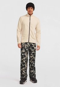 Bežová fleecová bunda s vysokým golierom, čierny zips, kombinovaná s nohavicami s čiernym a krémovým rozliatym vzorom a čiernymi topánkami.