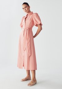 Roze, midi haljina na dugmad s puf rukavima, remenom u struku i glatkim materijalom. Nošena uz minimalističke sandale.