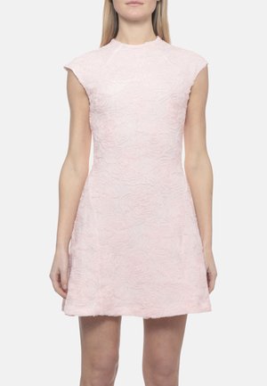 Donna con abito senza maniche di colore rosa pallido, tessuto e con motivi floreali e collo alto, in piedi con le braccia rilassate ai lati.