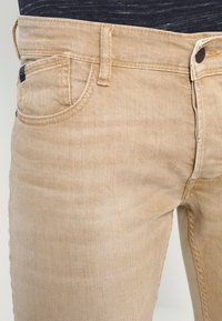 Jeans de mezclilla beige con corte ajustado, que cuentan con bolsillos delanteros, costuras cosidas y cierre de botón. La tela tiene una textura suave y ligeramente desgastada.