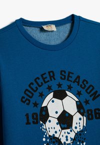 Niebieski bawełnowy sweatshirt z okrągłym dekoltem. Na przodzie znajduje się graficzny wzór piłki nożnej z napisem "SEZON PIŁKARSKI" oraz gwiazdami. Podano rok 1986.