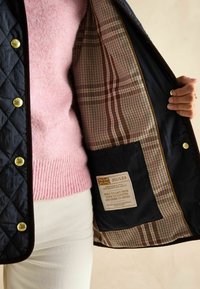 Granatowa pikowana kurtka z złotymi guzikami, z wnętrzem z materiału w kratę. Metka głosi "JOULES", z tekstem zapewniającym o trwałości i jakości.