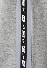 Šedá tkanina má vertikální černý pruh s opakujícím se bílým textem "NIKE". Textura je měkká a má hladký vzhled.
