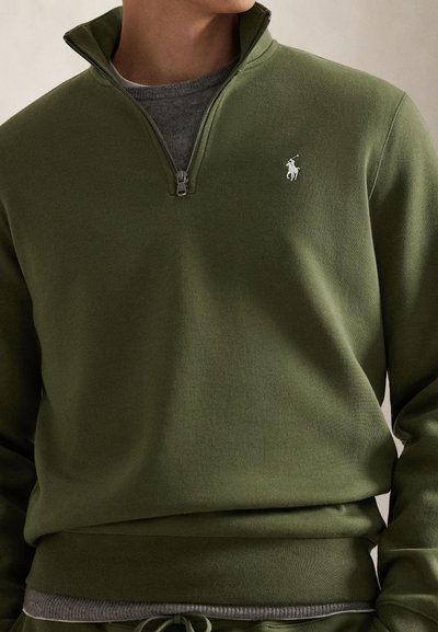 Πουλόβερ σε ελιά πράσινη απόχρωση με φερμουάρ μέχρι τη μέση, ριμπ τελείωμα και μανσέτες, με ένα μικρό λευκό λογότυπο στο αριστερό στήθος. Μαλακή υφ ή υφάσματος.