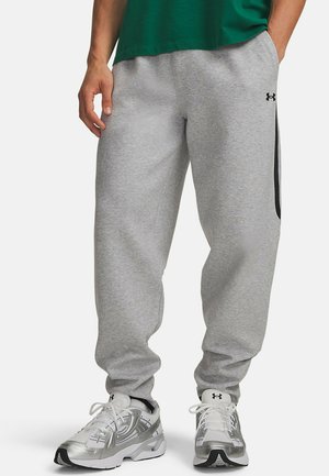 Person wears light grey Under Armour sweatpants, silver athletic shoes, and a green shirt. Hands rest at sides. Only lower body visible.