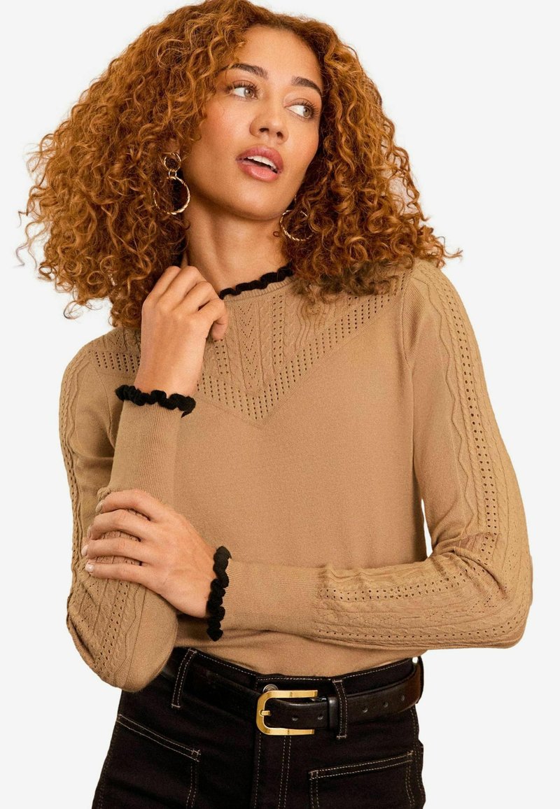 Maglione beige con design a trecce, dotato di un colletto e dei polsini ribattuti neri e arricciati. Il modello posa con le mani incrociate, indossando pantaloni neri.