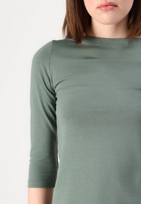 Grön, skräddarsydd topp med tre fjärdedelars ärmar, tillverkad av ett mjukt, slätt tyg. Urringningen är båtformad och texturen är lätt.