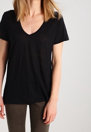 Vrouw met een zwart T-shirt met V-hals en korte mouwen en donkere, gestructureerde broek, staand tegen een effen lichte achtergrond.