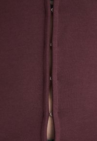 Top color bordeaux realizzato in tessuto morbido, caratterizzato da una spacco centrale con cuciture a contrasto e dettagli in metallo lungo l'apertura.