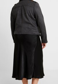 Giacca in similpelle nera con finitura opaca e spalle strutturate, indossata su una gonna nera in satin con una texture liscia e svasata.