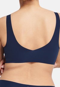 Mørkeblå bralette med brede stropper og dyp V-rygg. Stoffet har en subtil tekstur, som gir en sømløs og støttende passform.