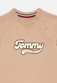 Sudadera beige con cuello redondo y mangas raglán. La parte frontal incluye "Tommy CLASS OF 1985" en letras grandes en negro y rojo.