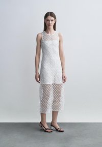 Žena v bielych midi šatách bez rukávov s čipkou a priehľadnou spodnou časťou, v čiernych sandáloch s blokovým opätkom na remienkoch, stojaca na šedom podlahe.