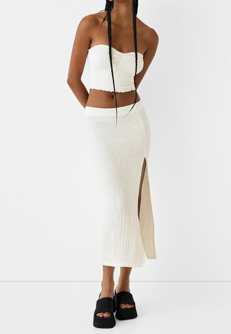Femme portant un haut court blanc texturé sans bretelles et une jupe assortie taille haute avec une fente sur la cuisse, associée à des sandales à plateforme noires.