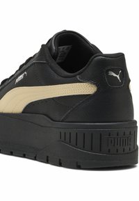 Sneaker nera in pelle con una striscia laterale beige, collo imbottito e suola nera testurizzata. Presenta il logo Puma sul tallone e sulla linguetta.