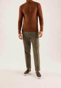 Homme portant un pull à col roulé en maille torsadée couleur rouille, un pantalon vert olive et des mocassins marron avec des semelles blanches, debout sur un fond clair.