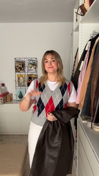 Gilet en laine gris avec un motif en losanges rouges et bleus, t-shirt blanc, et tenant une veste en cuir marron foncé. Vêtements disposés en arrière-plan.