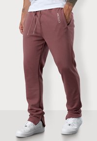 Bordeauxrote Jogginghosen mit elastischem Bund, Seitentaschen und dem Markennamen "ESTEEM" in Weiß aufgedruckt, kombiniert mit weißen Turnschuhen.