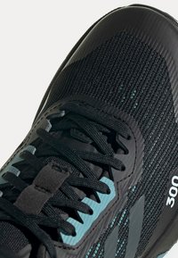 Scarpa sportiva nera con tomaia testurizzata, caratterizzata da accenti turchesi, lacci neri e un logo del marchio ben visibile sul lato.