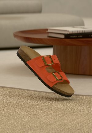 Oranžinis sandalų modelis su dviem dirželiais, auksinėmis sagtimis ir kamštine padu, sklendžiantis virš austo smėlio spalvos kilimėlio, medinio apvalaus stalo ir knygų fone.