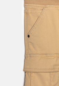 Béžové cargo kalhoty mají velkou boční kapsu, zesílené švy a elastický pas. Materiál se zdá být odolný s hladkým povrchem.