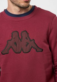 Bordo sweatsirt iz mehkega blaga z črnim vezenim logotipom dveh sedečih figur. Ovratnik v stilu posadke in rebrasti manšeti.
