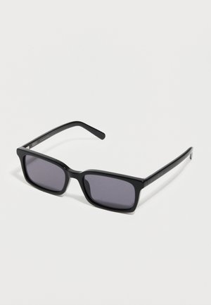 Lunettes de soleil rectangulaires noires avec des verres teintés foncés posées sur une surface blanche, légèrement inclinées vers la gauche.