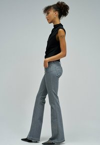 Kvinna med lockigt hår i en svart ärmlös topp och grå utsvängda jeans, stående i profil med händerna i fickorna mot en enkel bakgrund.