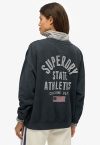 Felpa blu navy scuro con vestibilità oversize, con la scritta "Superdry State Athletics" stampata sul retro e dettagli a righe sul colletto. Materiale di cotone.