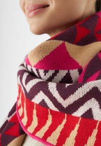 Frau lächelt sanft, trägt einen bunten gestrickten Schal mit roten, rosa, beigen und lilanen geometrischen Mustern, der um ihren Hals gewickelt ist.