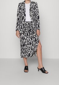 Ensemble blazer et jupe à imprimé léopard noir et blanc, avec une fente haute sur la jupe et des mules ouvertes noires. Texture lisse.