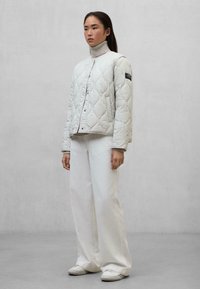 Gequiltede witte puffer jacket met een hoge kraag en ribbelmanchetten, gecombineerd met losse witte broek en witte instapschoenen, staande tegen een grijze achtergrond.