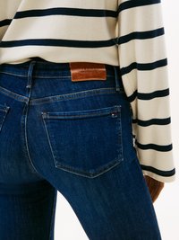 Gros plan sur une personne portant un jean en denim bleu foncé et une chemise à manches longues crème avec des rayures horizontales noires, montrant la poche arrière et l'étiquette de la ceinture.