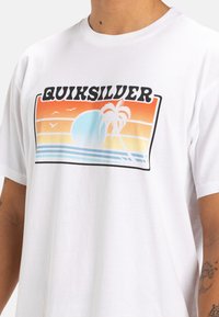 T-shirt bianco in cotone con una grafica colorata che rappresenta un tramonto, una palma, il sole e onde oceaniche, con il marchio "Quiksilver" nella parte superiore.