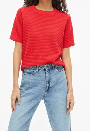 Vrouw die een gestructureerde, felrode, kortärmelig gebreide top draagt, ingestopt in lichtblauwe high-waisted denim jeans met een ontspannen pasvorm.