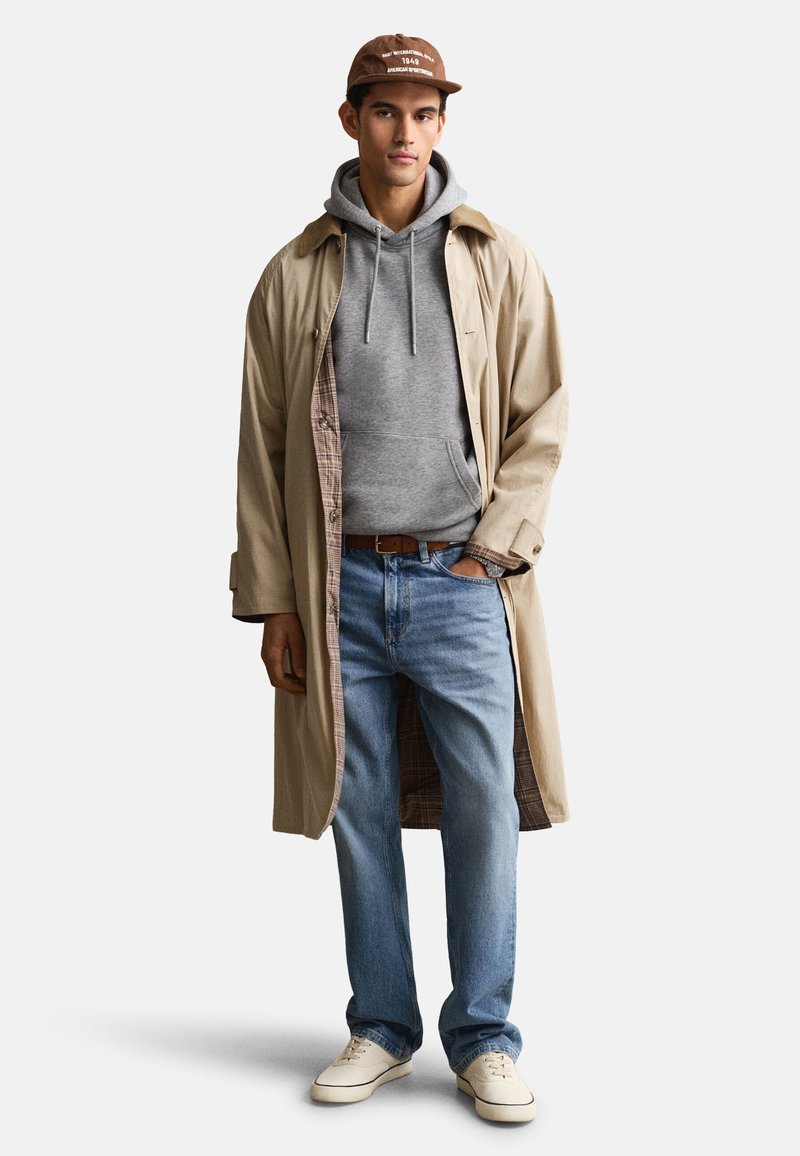 Joven con gorra marrón, abrigo largo beige, sudadera con capucha gris, vaqueros azules y zapatillas blancas, de pie con una mano en el bolsillo.