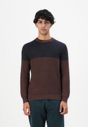 Junger Mann in einem gerippten, langärmligen Pullover mit marineblauen und braunen Farbsprenkel, vor einem schlichten weißen Hintergrund stehend.