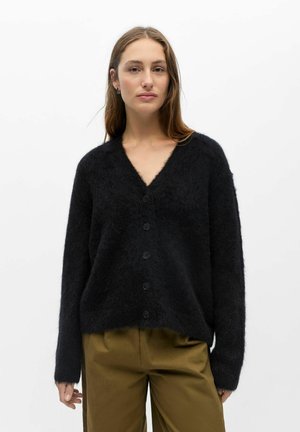 Femme aux longs cheveux portant un cardigan noir duveteux boutonné et un pantalon vert olive sur un fond blanc uni.
