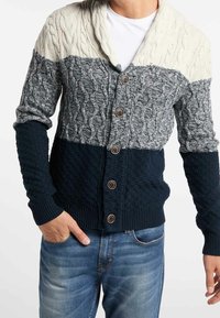 Cárdigan de punto con cuello chal; presenta bloques en crema, gris y azul marino, textura de punto trenzado y botones marrones en la parte delantera.