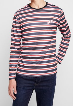 Homme portant un t-shirt à manches longues à rayures horizontales bleu marine, blanc et rouge avec le texte « Good » sur la poitrine et un jean bleu foncé.
