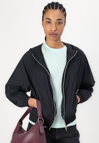 Giacca bomber nera in nylon con cappuccio e zip dorata, indossata sopra una maglietta leggera color menta. Il modello tiene in mano una borsa in pelle bordeaux.