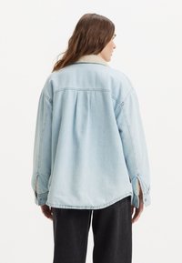 Lys blå denimjakke med en koselig shearling-krage, senkede skuldre og en avslappet, løs passform med sidespalter i sømmen.